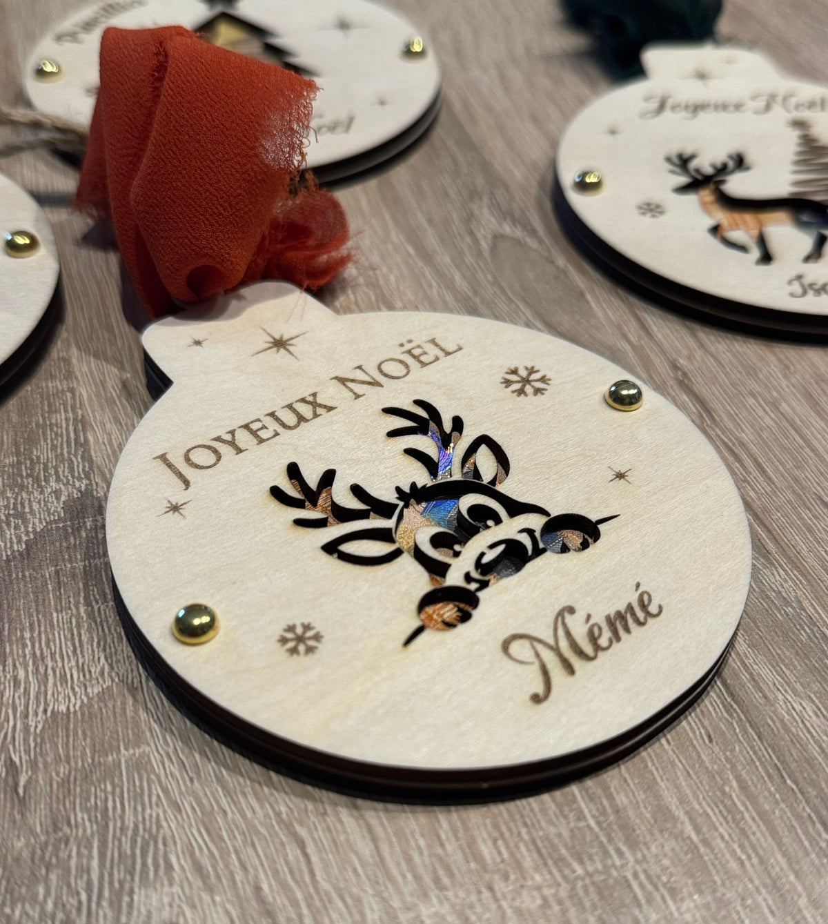 Boule de Noël en bois personnalisée « Joyeux Noël Mémé » avec bébé renne découpé laissant voir un billet, flocons gravés et ruban terracotta